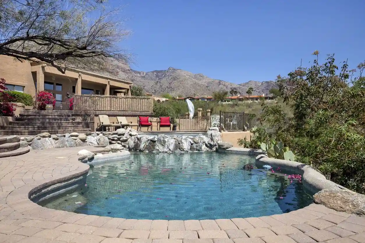 A backyard swimming pool by Super Clean Pools with a stone border and small rock waterfall feature, surrounded by desert landscaping and a patio area with outdoor seating; mountains visible in the background under a clear blue sky.