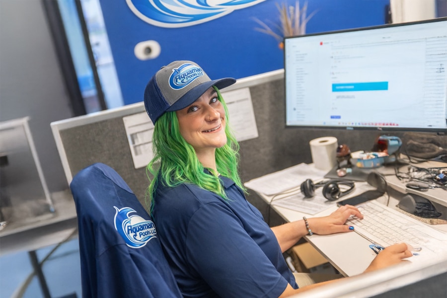 Person with green hair wearing a blue Aquarama Pools shirt and hat sits at a desk, smiling at the camera, with a computer monitor, keyboard, and various office items visible in the workspace.