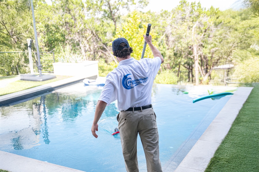A person wearing a white shirt and khaki pants uses a pool skimmer to clean a backyard swimming pool surrounded by greenery on a sunny day.