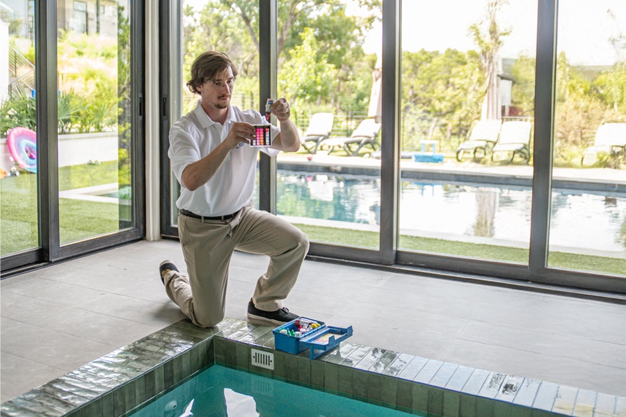 A man in a white shirt kneels by an indoor pool, holding a water testing kit. The kit is open on the pool edge. Large windows reveal an outdoor pool and lounge chairs.